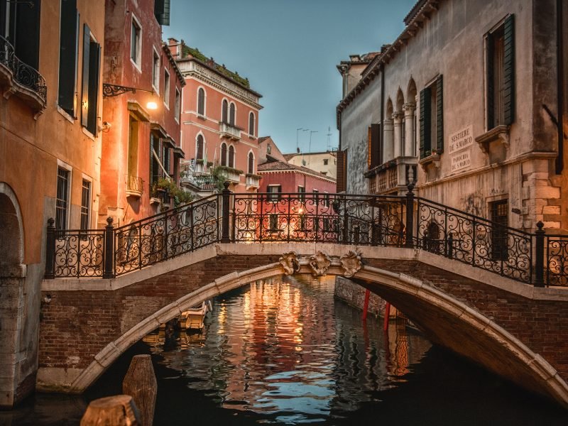 Blue hour lighting going through the canals and bridges of Venice, down a small nondescript alleyway with an arched brick bridge that bridges the two canals.