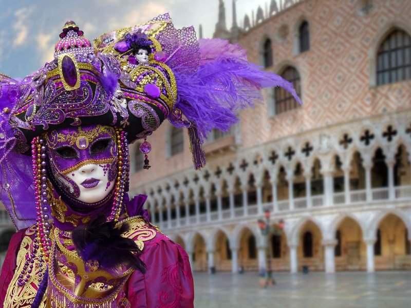 Feminine looking person in a Venice Carnival Mask with purple embellishment standing in an empty St Marks Square, celebrating Carnival in Venice in February.