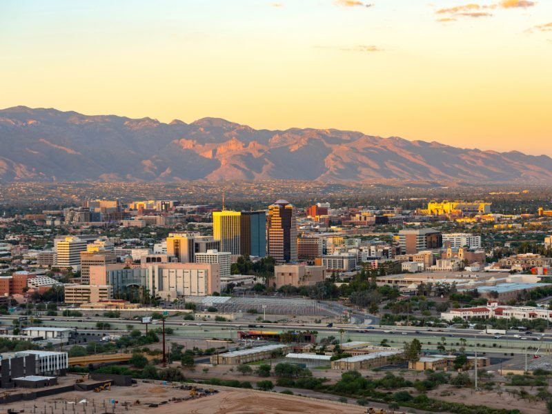The skyline of Tucson at sunset with mountains in the distance and buildings in the foreground.