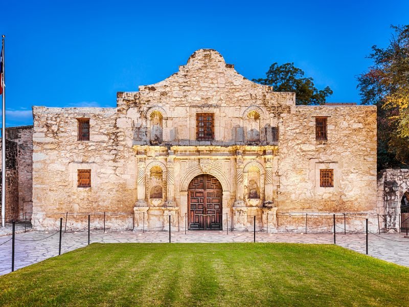 The front facade of the Alamo, a historic battle site in Texas, with a green lawn in front and dark sky