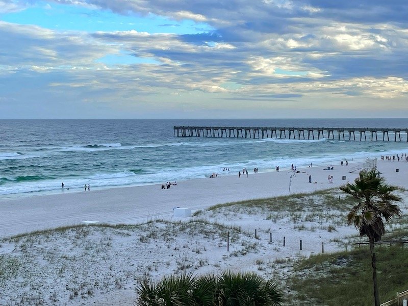 The pier leading out into the ocean in Pensacola FL on a cloudy day with a handful of people lounging on the beach at sunset time