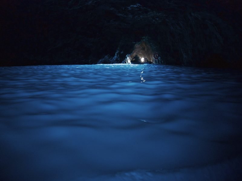 Another photo of the interior of the Blue Grotto sea cave in Capri. The water is glowing turquoise and you can see a tiny boat near the entrance which looks just like a tiny pinprick of light coming through.