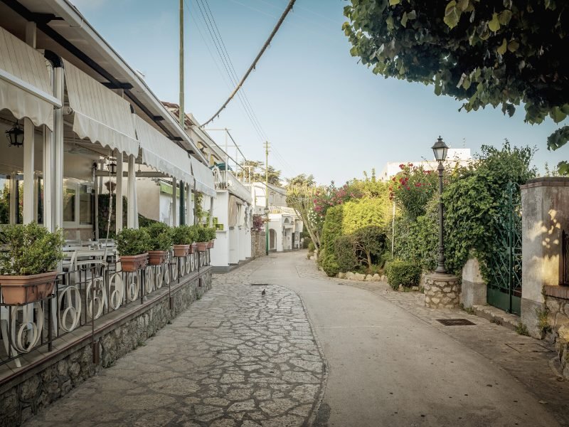A quiet street scene in the town of Anacapri, with a small restaurant with no one in it and greenery and trees.