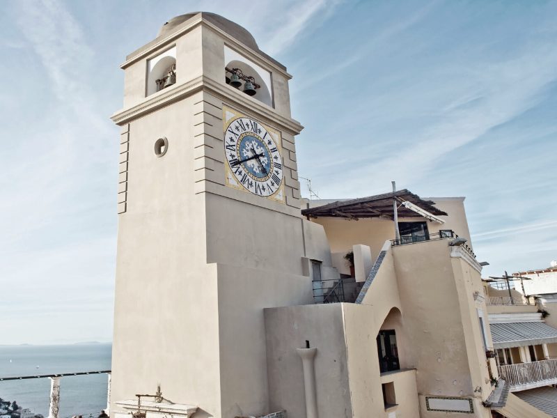 Famous clock tower in the Piazzetta of Capri with a tile clock face and sand-colored building around it.
