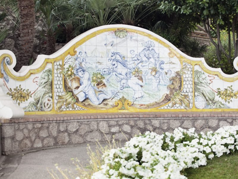 Some beautiful telework forming a bench in the Gardens of Augustus in Capri, with some white flowers in the foreground, and a footpath leading to more of the gardens.