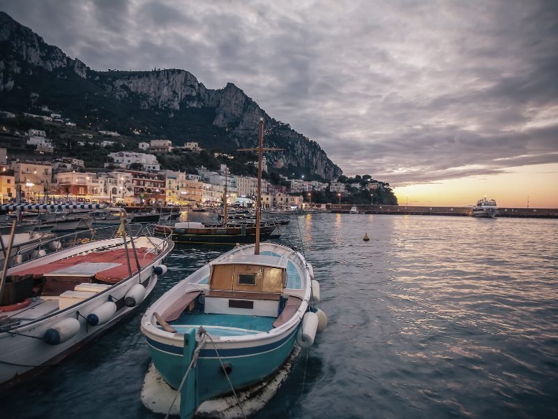 Boats in the water at Marina Grande in Capri with lit up buildings and a ferry in the distance, sunset approaching