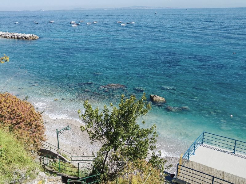 Rocky sandy beach with blueish turquoise water with boats and people enjoying the water on a summer day