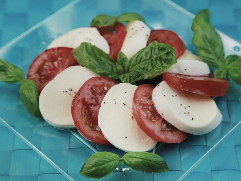 Caprese salaad with slices of tomato and mozzarella and basil on a clear plate on a blue placemat