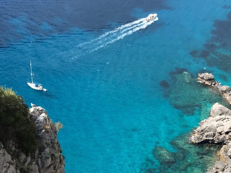 Two boats on the brilliant crystalline waters of Capri, looking see-through so that you can see the sea bottom, with wake trails from the boats on a brilliant summer day.