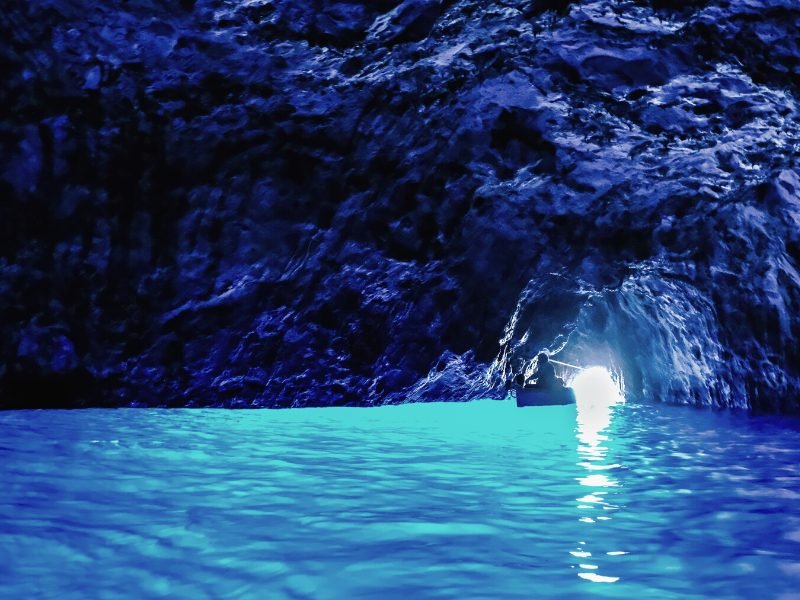 A very iridescent and bright blue interior shot of the Blue Grotto, with one boat visible near the cave entrance, and the light makes the water look like it is glowing and turquoise.