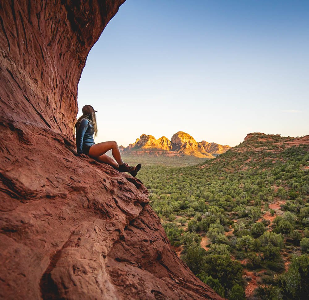 The author, Jessica, sitting on a red rock in Sedona looking away towards the light on the red rocks in the distance, wearing hiking clothes and shorts and a hat.