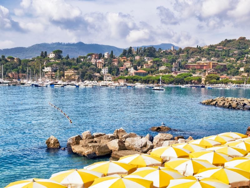 yellow and white umbrellas on the scenic shoreline of the Italian riviera with buildings on the land and sailboats in the water