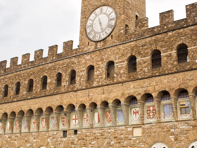 Several crests in the archway windows of the Palazzo Vecchio, with other windows and a large clock face with Roman numeral numbers, building made of stucco-colored brick.