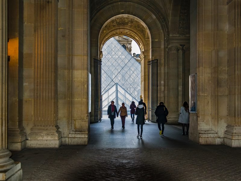 People in winter clothing visiting the Louvre, in a series of arches from which you can see the glass pyramids on the other end.