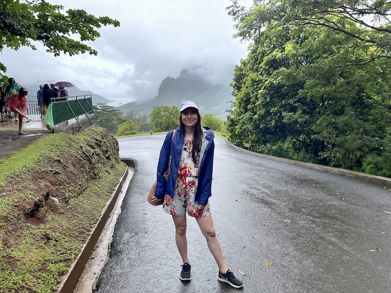 Allison wearing a navy rain jacket, floral romper, and white baseball cap on a rainy day at an overlook in Moorea.