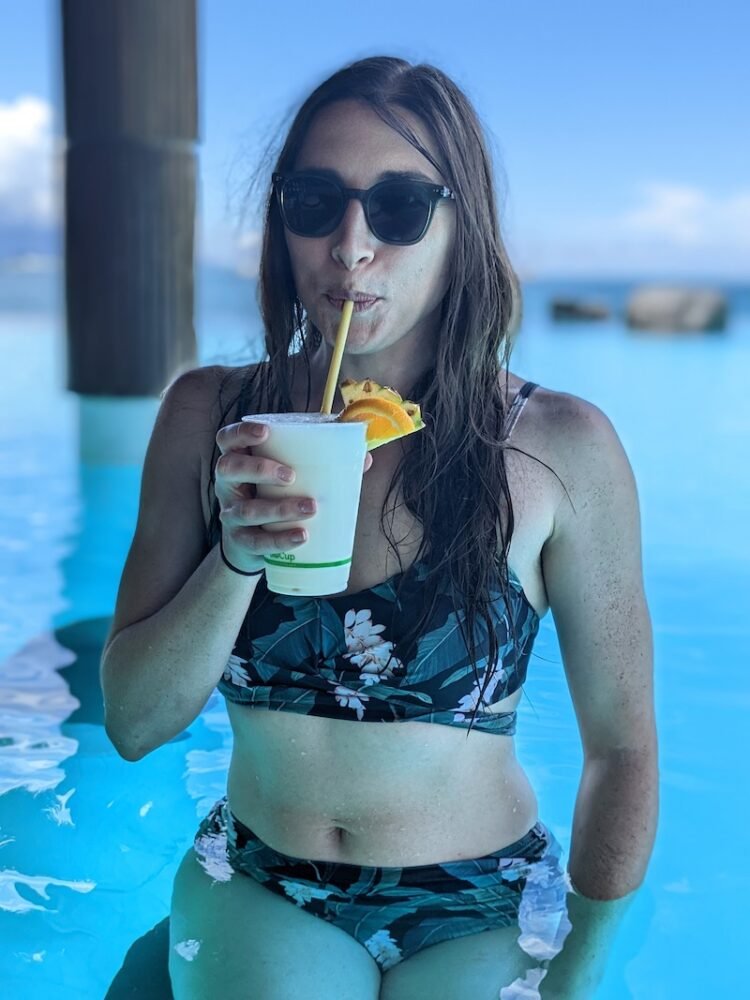 Allison sipping a virgin pina colada while wearing sunglasses in a monstera-print bikini with a high waist with blurry pool in background