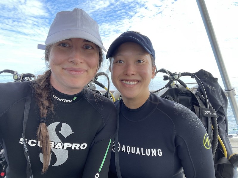 Allison Green and her partner wearing scuba gear on a boat. Allison is wearing a Scubapro wetsuit and her partner is wearing an Aqualung wetsuit. They are both smiling at the camera and wearing hats with their hair back and scuba gear visible behind them.