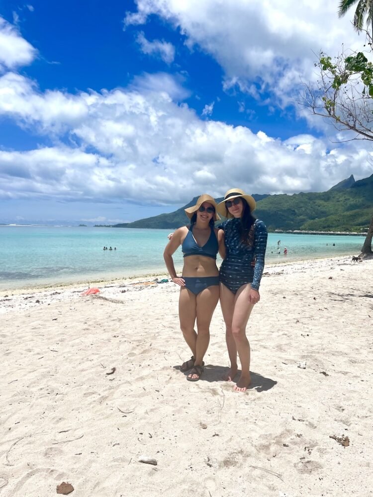 Allison and her partner wearing sunhats and swimsuits on the sandy beach on a lagoon of Moorea with bungalows in the distance background