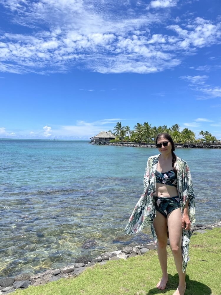 Allison in a bathing suit and flower-print cover up, sunglasses and hair in braid, with coral reef, lagoon, and bungalows in the distance.