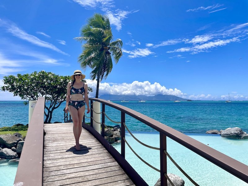 Allison exploring Tahiti while staying at a resort, wearing a bikini, with infinity pool, palm tree, blue sky, and Moorea in distance, while walking barefoot on a bridge.