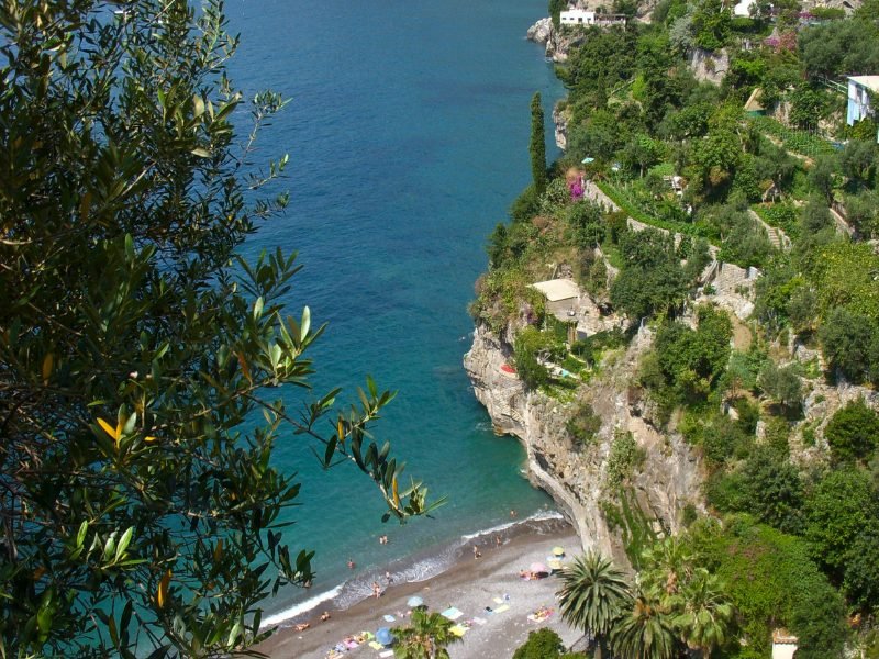 View from above the steep cliff over Arienzo Beach, with terraced cliffs with gardens and groves built into the landscape, with a beach below with deep blue water and sunbathers enjoying the quiet beach on the Amalfi Coast