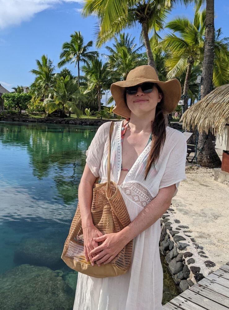 Allison holding a tan mesh beach bag at a resort in Tahiti, packing list essential item!