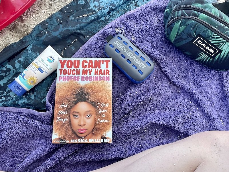 Purple beach towel with sunscreen, book called "You can't touch by hair" by Phoebe Robinson, fanny pack, and travel towel