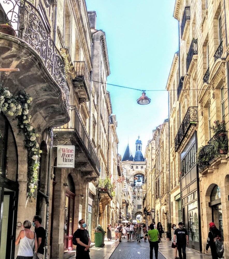 People in the street of Bordeaux, many wearing masks in the post pandemic era, a bell tower and church in the distance, wine shops and stores around