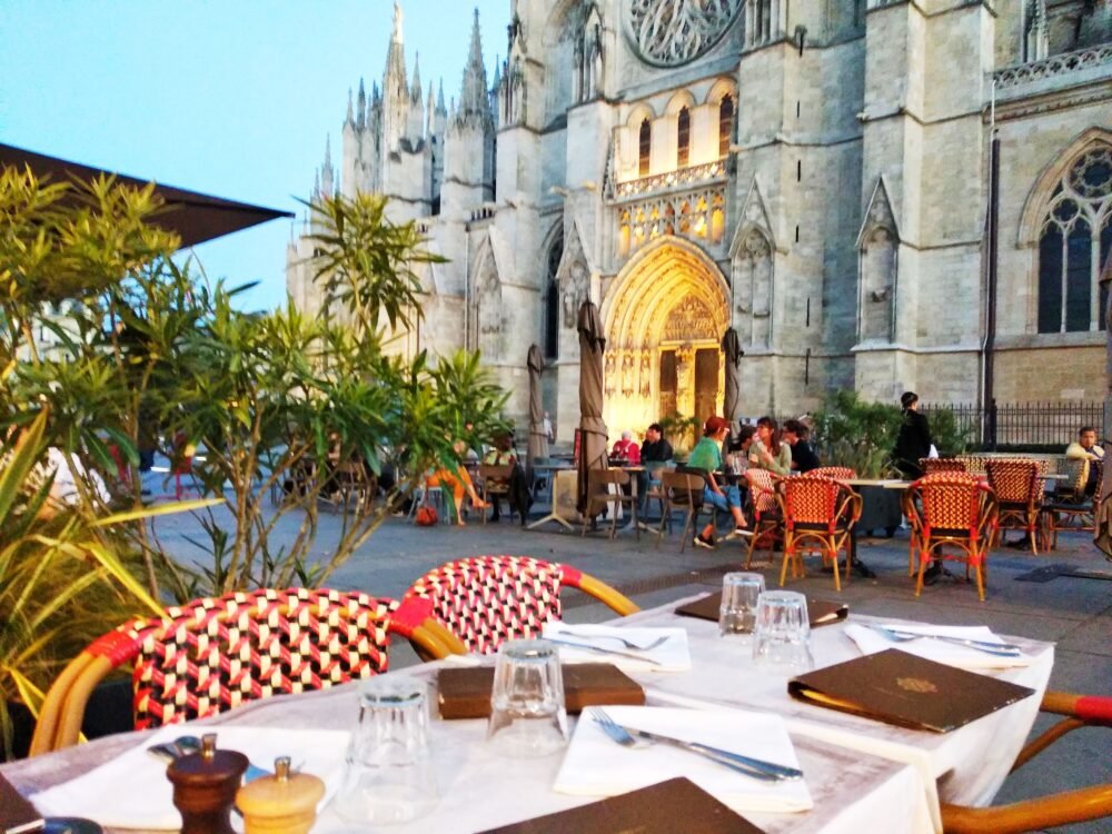 Bistro style chairs outside dining al fresco in the blue hour with soft light and lights coming on in the church behind, a few people already eating in the early evening