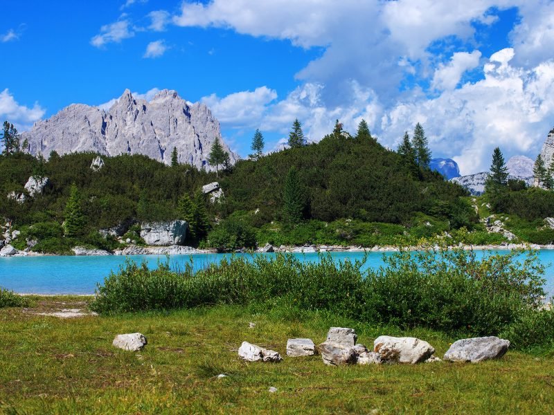 View of turquoise waters of Lake Sorapis with trees, rocks, glassy still water, mountain peaks, clouds in the sky on a beautiful day