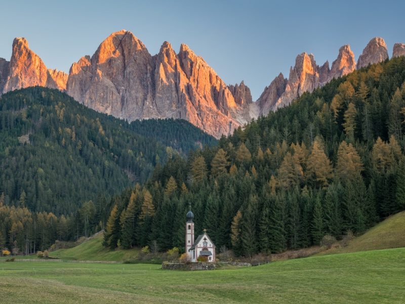 Beautiful church in the Dolomites, called the Church of San Giovanni in Ranui, located in a valley at the base of many mountain peaks that are lit up with golden afternoon light.
