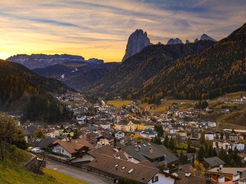 Town in the valley down below, small town of Ortisei surrounded by mountain peaks and trees in yellow and green colors