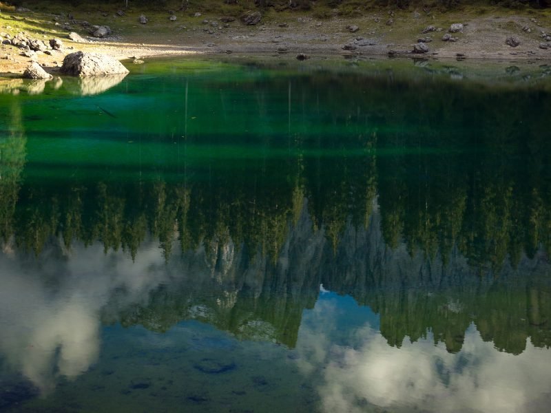 Reflection in the lake of Lago di Carezza, showing the mountains reflecting in the still water and the pine trees and the clouds