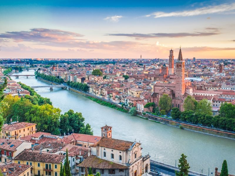 Verona city center with river flowing through it at sunset, with 3 bridges crossing the river, and views of church towers and campaniles in the skyline on the far side of the river.