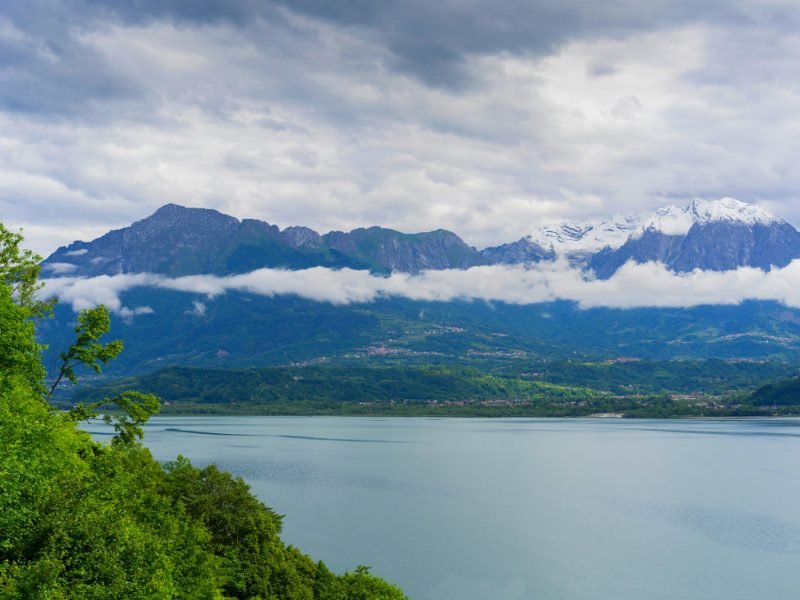 Beautiful alpine lake named Lago di Santa Croce, part of the Veneto region of Italy, with small towns dotting the shore of the lake and clouds and some snow on the peaks of faraway mountains