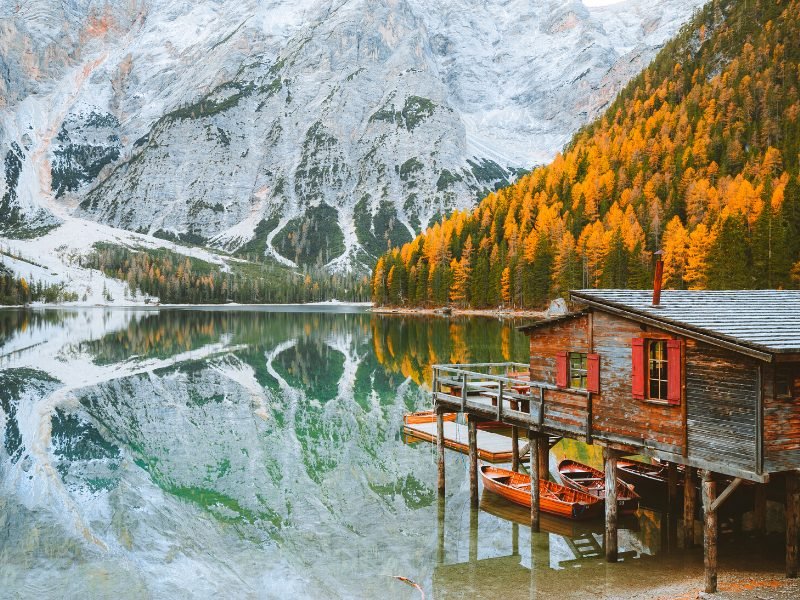 Fall scene in the Dolomites with lake reflecting boathouse and trees and mountain background like a mirror, with red boathouse and yellow trees on the right side of the picture