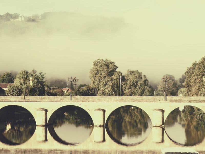 View of the Vezere river, with arched bridge making a pathway across, reflecting the bridge, and fog and town on the hill above