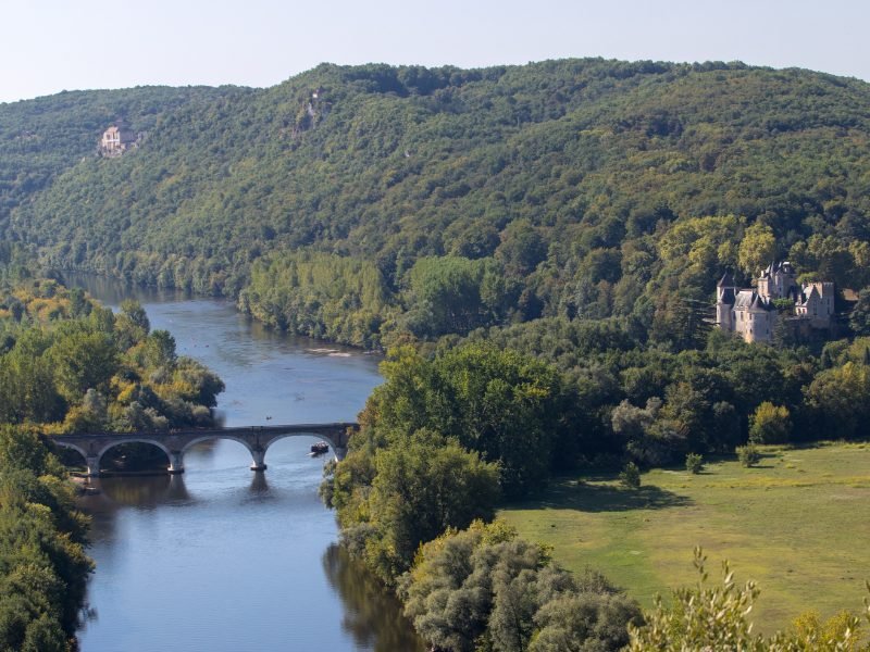 A river with a bridge and a boat, blue sky, forest around it with a large castle and an open grassy space in the Dordogne region of France