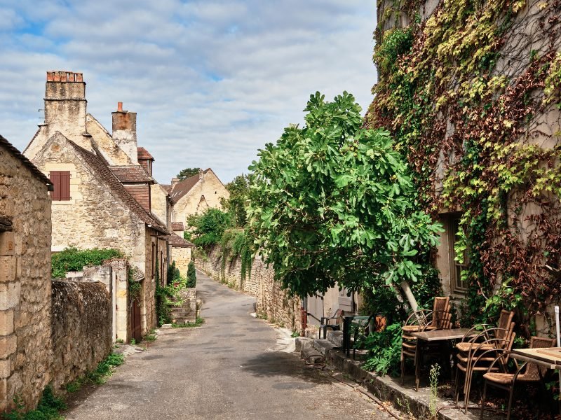 A narrow alleyway in Domme, France, with stacked chairs, medieval houses, and trees and ivy growing on one of the walls