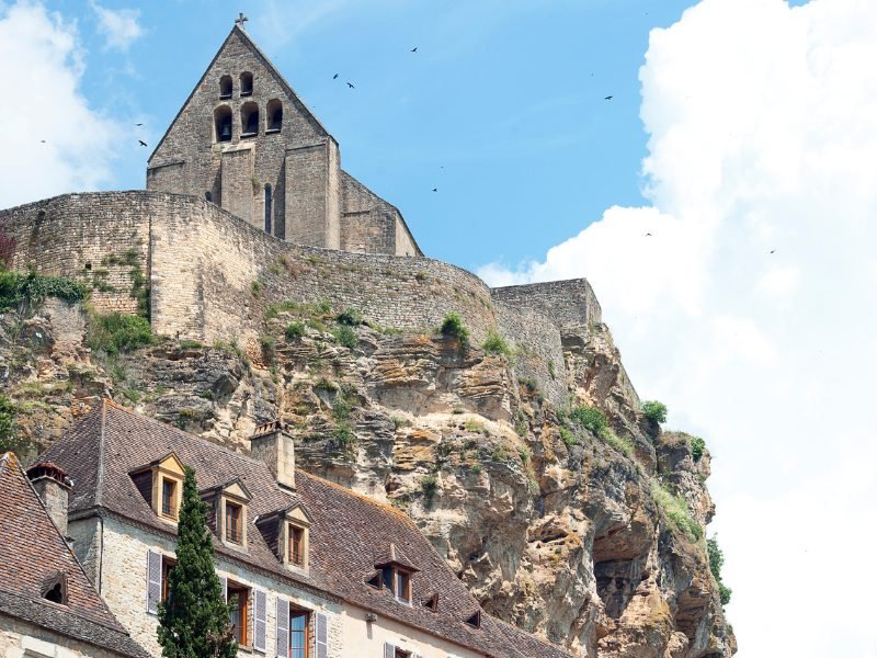 A church on top of a large rocky outcrop, overlooking a historic and cute Dordogne village, on a partly cloudy day with birds in the sky