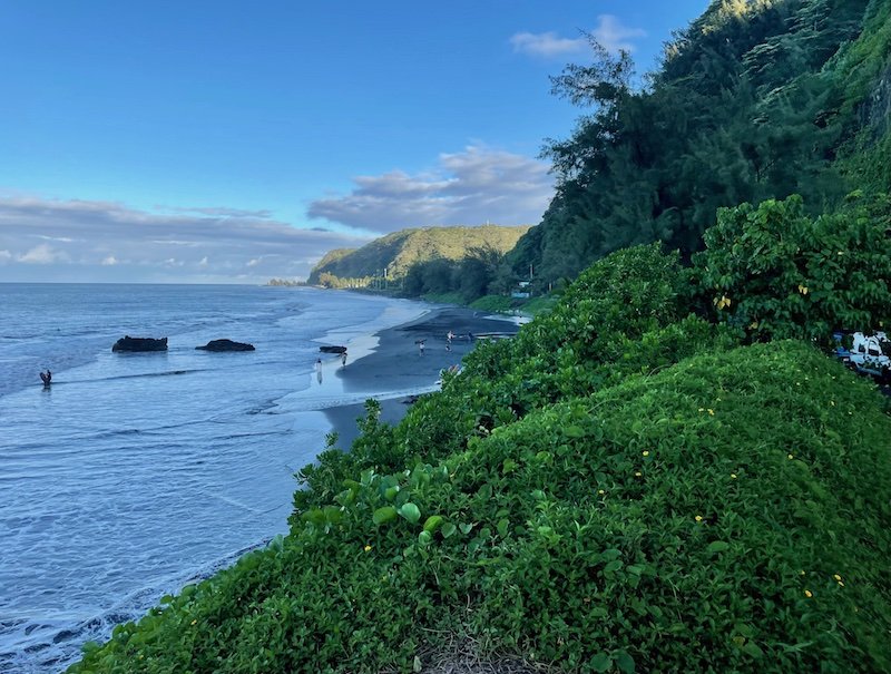 Black sand beach with people bringing their surfboards out, and curvy road on the coastline