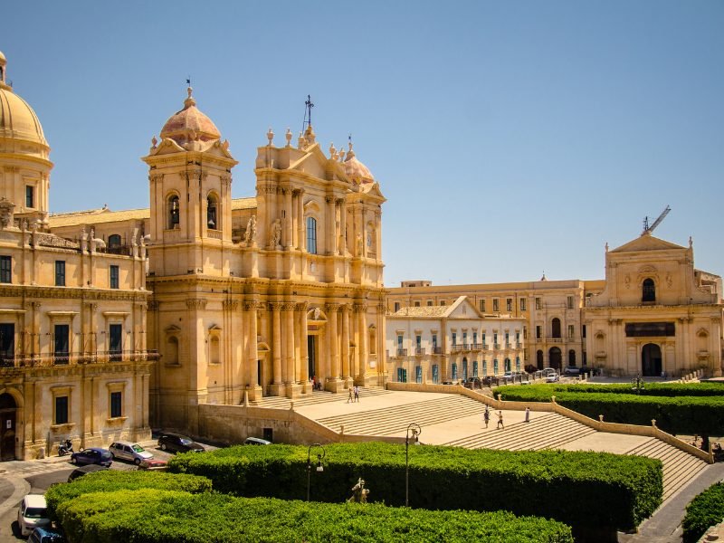 The famous cathedral in Noto Sicily, with grounds and stairs and a few people walking towards it on a sunny day.