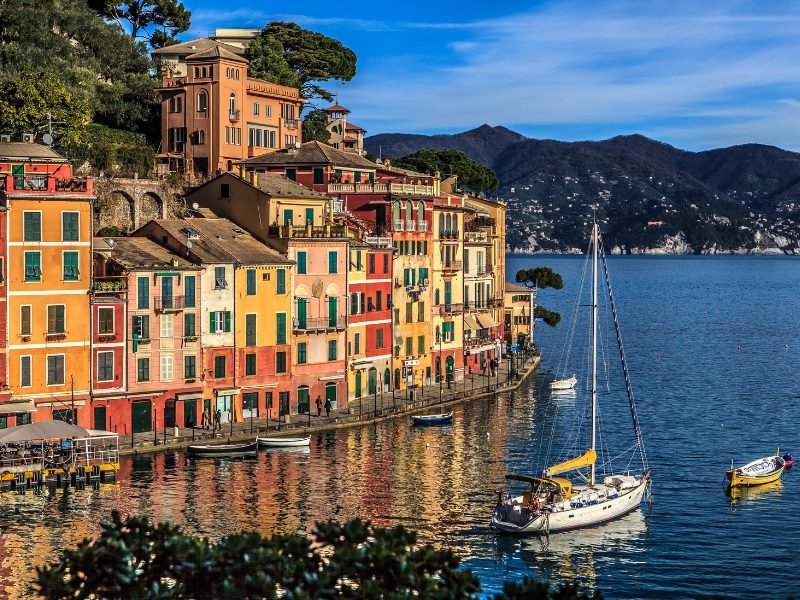 The colorful fishing village of Portofino on the Ligurian coast with dark blue water, sailboat, and smaller canoe-style boats in the water
