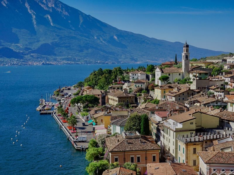 Dark blue waters of Lake Garda with lakeside town with beautiful buildings, church tower, and mountains in background