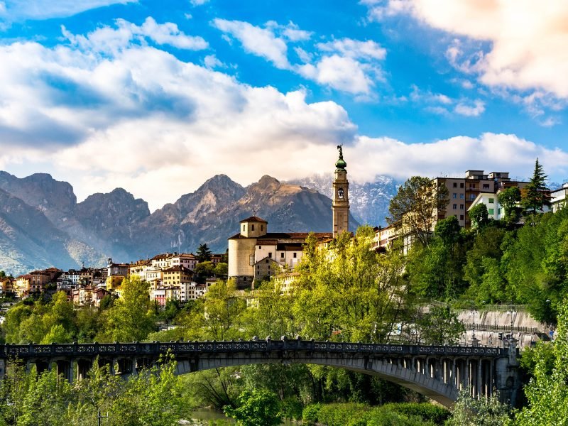 The late afternoon sunlight on the mountains of the Dolomites and the charming town of Belllno, with a church tower and a bridge visible in the framing of the town