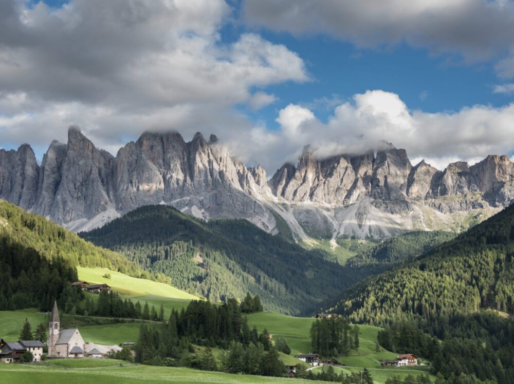 the rugged peaks of the dolomites with grassy foothills with trees, churches, small towns at the foot of the majestic peaks, with clouds in the sky