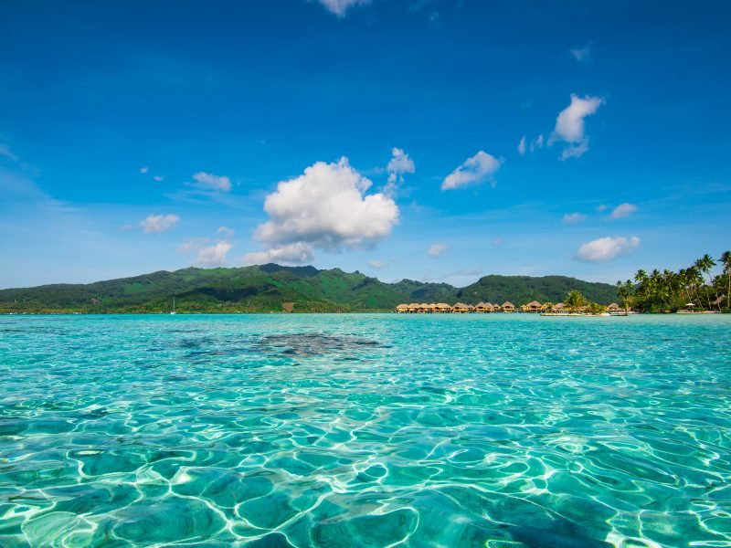 Crystal clear water in the lagoon with Tahiti overwater bungalows of Le Taha'a island visible in the distance, with green island on horizon as well.