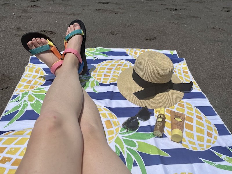 Pineapple print microfiber towel with sunscreen products and sunhat and legs with rainbow sandals on a black sand beach