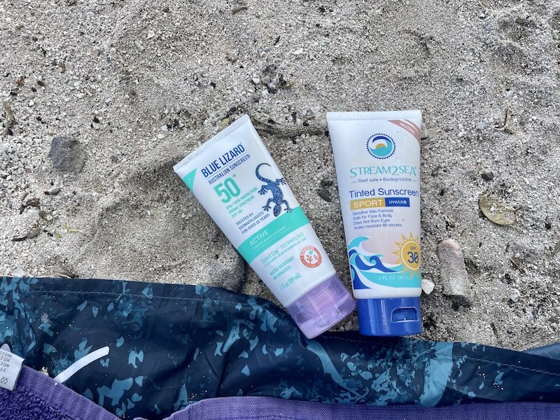 A side by side comparison of two reef safe sunscreen companies on a beach towel on the beach in Moorea