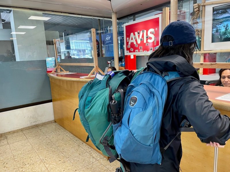 Woman with two backpacks at the Avis counter discussing with a rental car agent